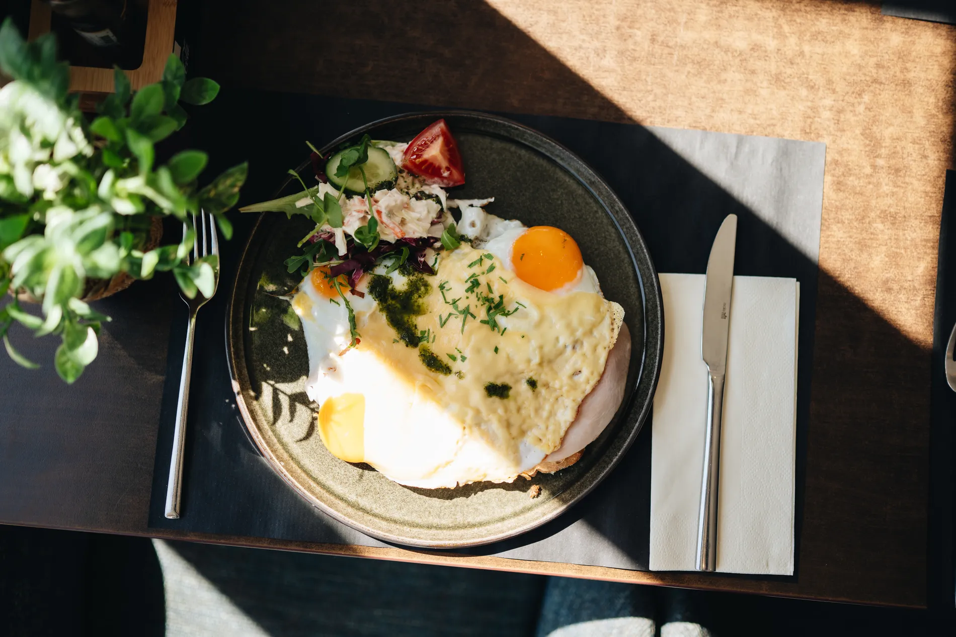 Bovenaanzicht van een uitsmijter op brood met spiegeleieren, ham, gesmolten kaas en pesto, netjes gepresenteerd met een salade van komkommer, tomaat en dressing.